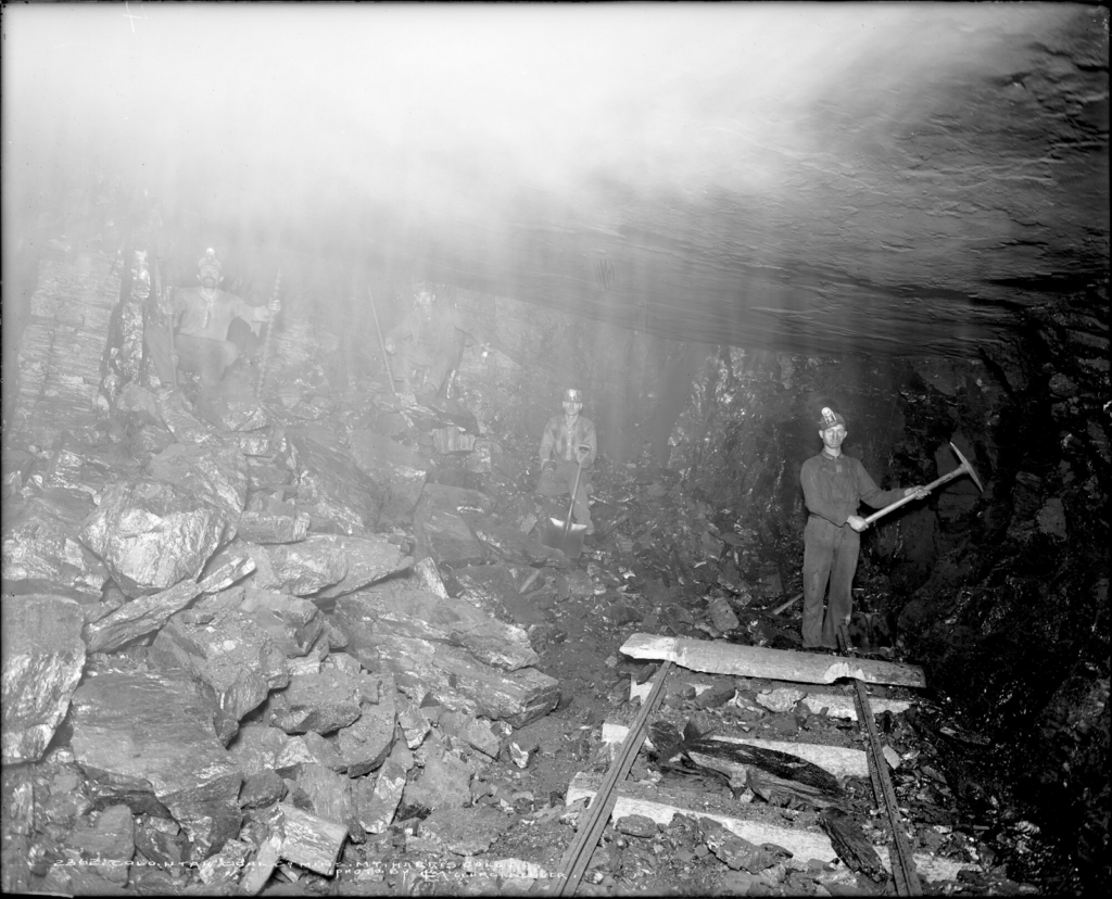 Interior view of a coal mine owned by the Colorado-Utah Coal Company at Mt. Harris, Colorado; at right, the rail line terminates and a miner stands near it, holding a pickax; at left, three miners sit; one holds a shovel, the others hold drills.