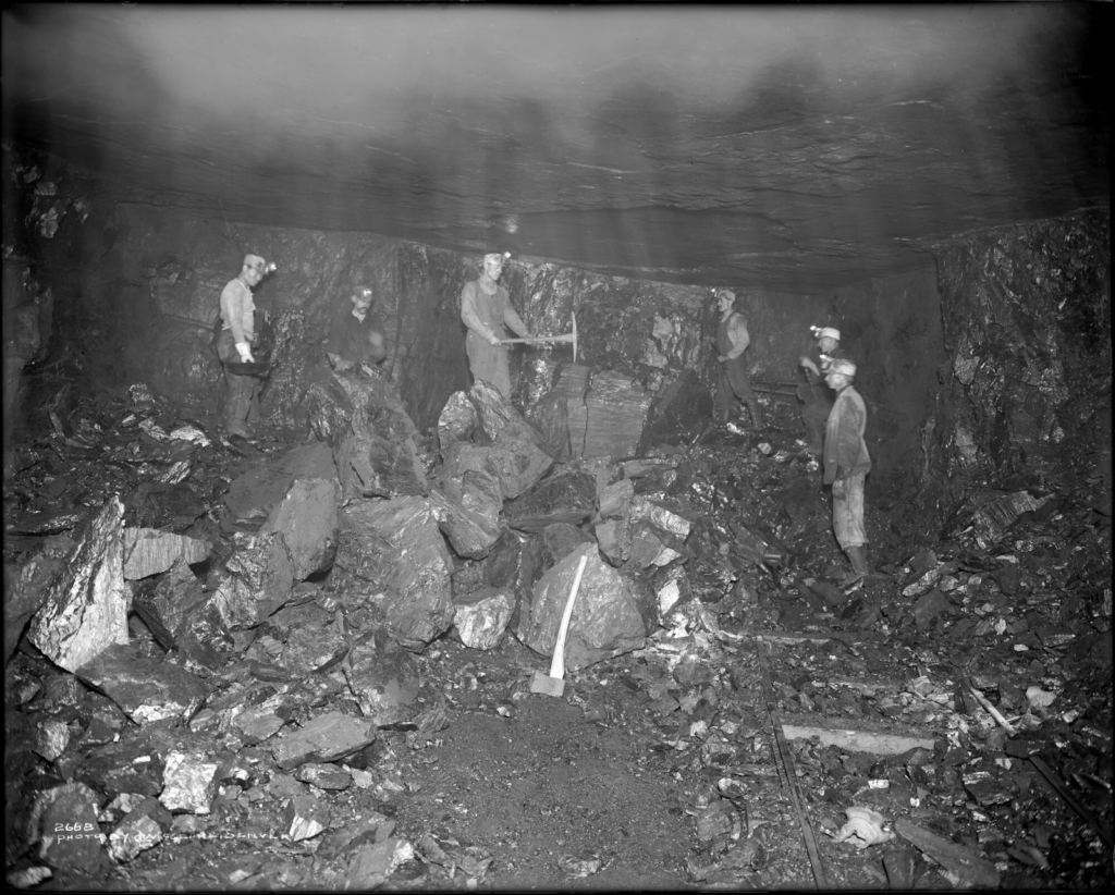 View of five miners in an underground coal mine owned by the Colorado-Utah Coal Company at Mt. Harris, Colorado; shows miners holding drills and pickaxes, a hatchet propped against a rock, and trolley tracks.