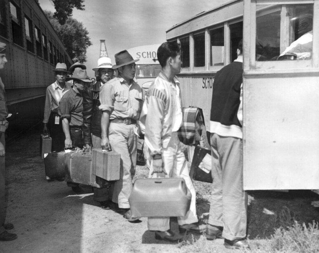 Japanese men arrive from the Merced Assembly Center, California, by train, carrying suitcases and boarding school buses on their way to Camp Amache, Granada Relocation Center, Prowers County, southeastern Colorado. A United States soldier in uniform stands next to the passenger train car and looks on as the line of evacuees board the bus. August 28, 1942.