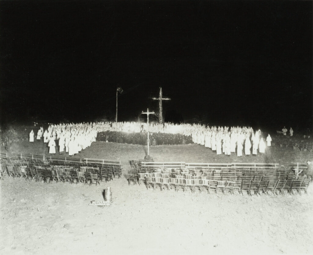 A cross burns at a night meeting of the Ku Klux Klan (KKK) on Table Top Mountain near Golden (Jefferson County), Colorado. Men in white robes and hoods encircle a group of men in street clothes who kneel in front of the burning cross. A container and cups on a tray sit behind rows of chairs.