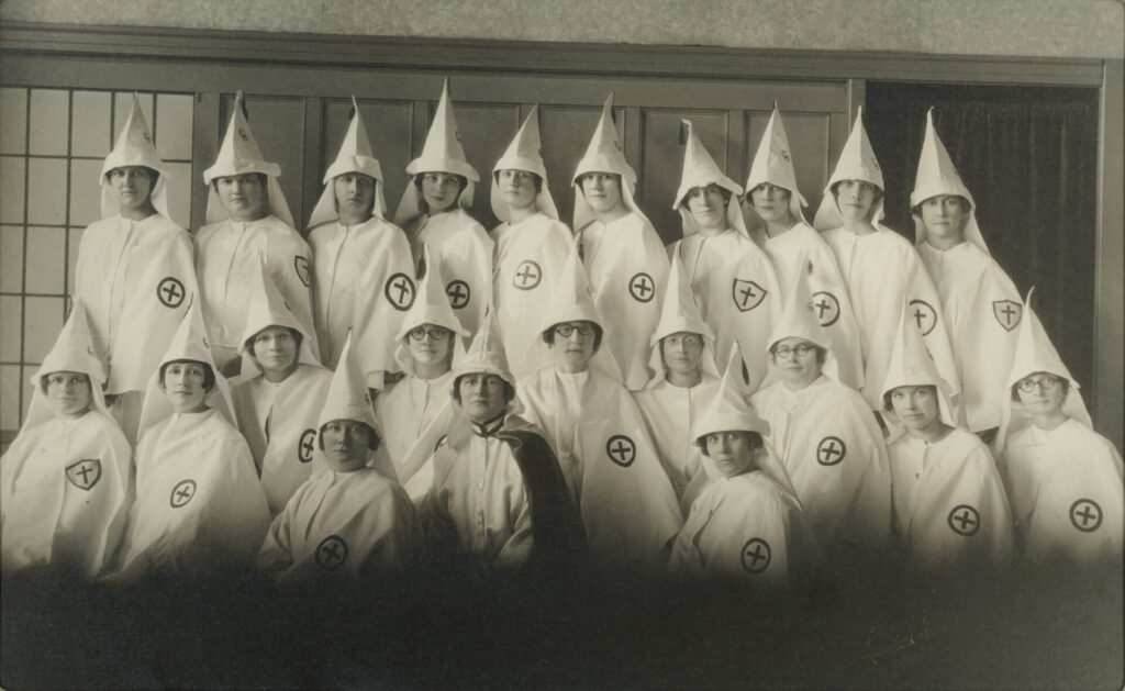 Studio portrait of women members of the Ku Klux Klan (KKK). The women pose in white hoods and robes with cross emblems. One woman wears a dark cape from 1922-1925
